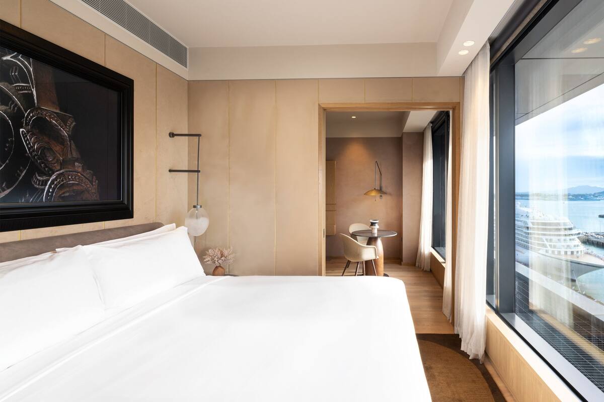 Ein Hotelzimmer mit einem großen Bett, einem Esstisch mit zwei Stühlen, einem kleinen Tisch mit einer Vase und einem großen Fenster mit Blick auf ein Kreuzfahrtschiff und den Hafen.