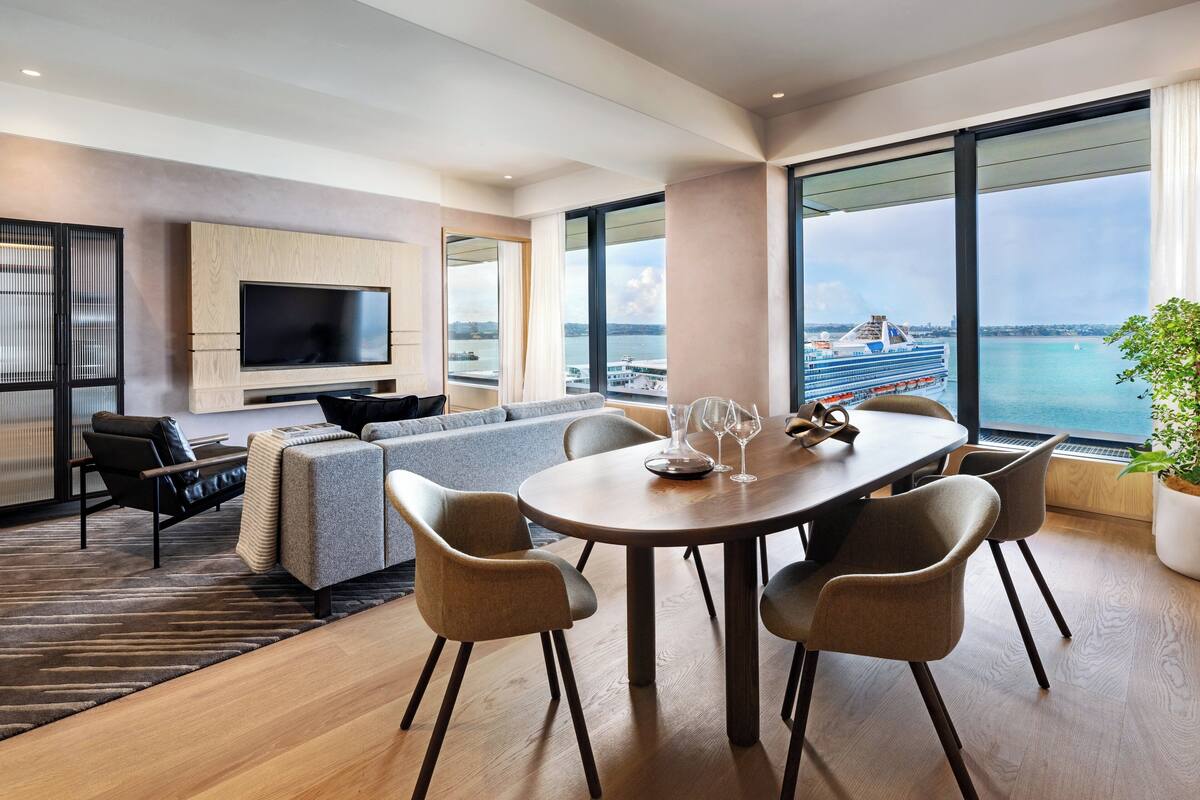 Ein modernes Wohnzimmer mit großem Fernseher, einem Esstisch mit Stühlen, einer Couch und Blick auf ein Kreuzfahrtschiff.