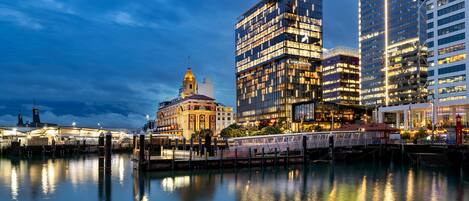 A waterfront cityscape with modern buildings, a historic building with a clock tower, and a bridge over the water.