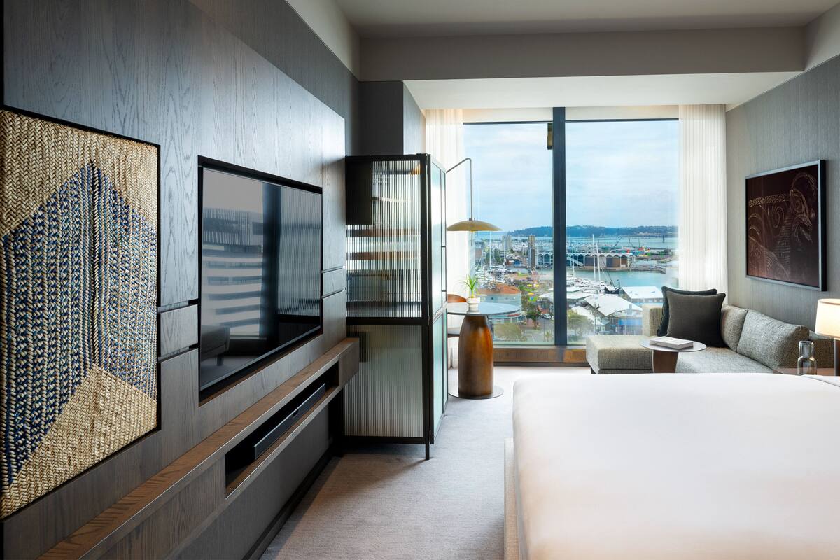 Ein modernes Hotelzimmer mit einem großen Bett, einem Fernseher, einer Sitzecke mit Stadtblick und einem Balkon mit Blick auf den Hafen.