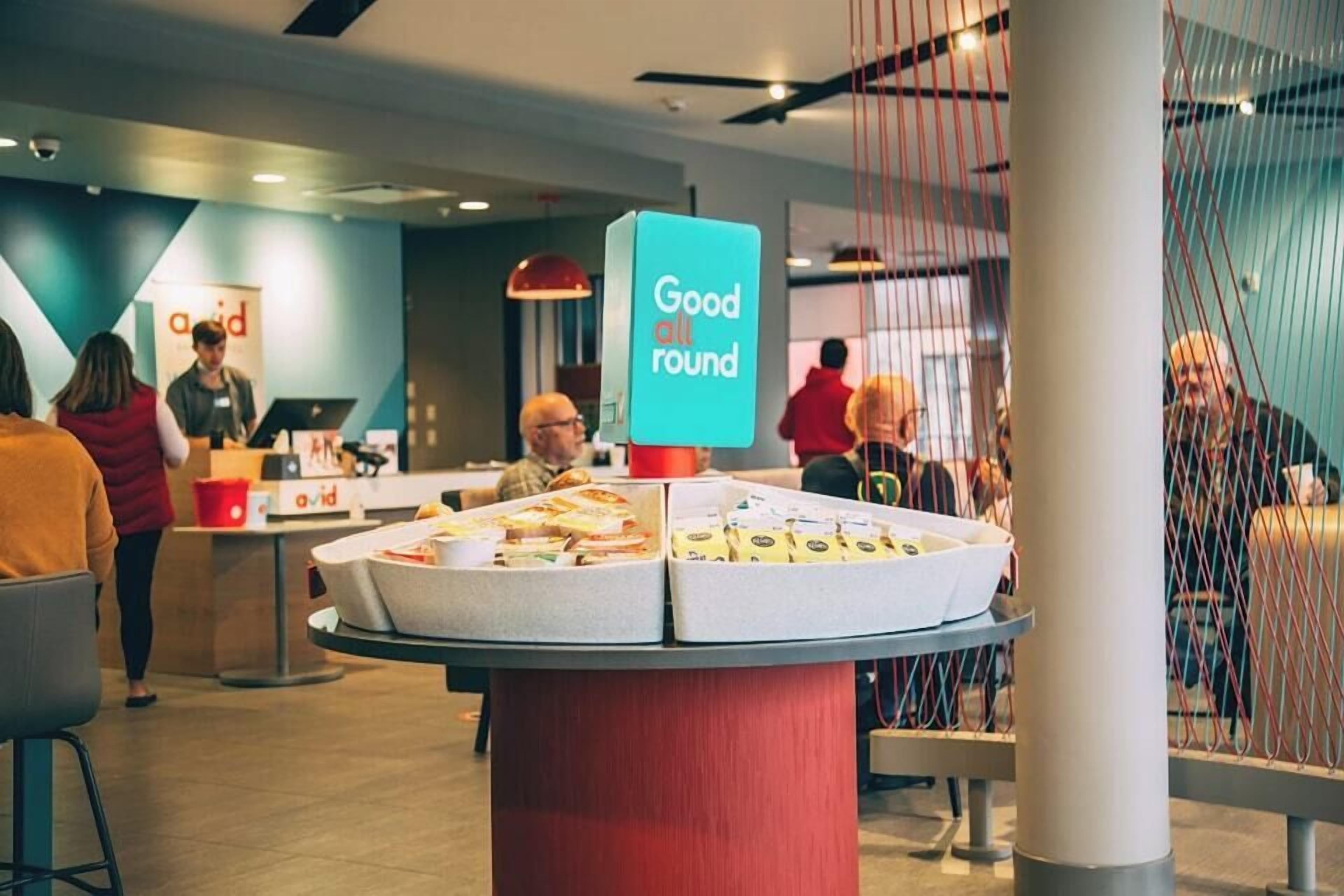 Um espaço interior moderno com uma mesa central exibindo alimentos, um letreiro com a inscrição 'Ótimo em tudo' e pessoas ao fundo.