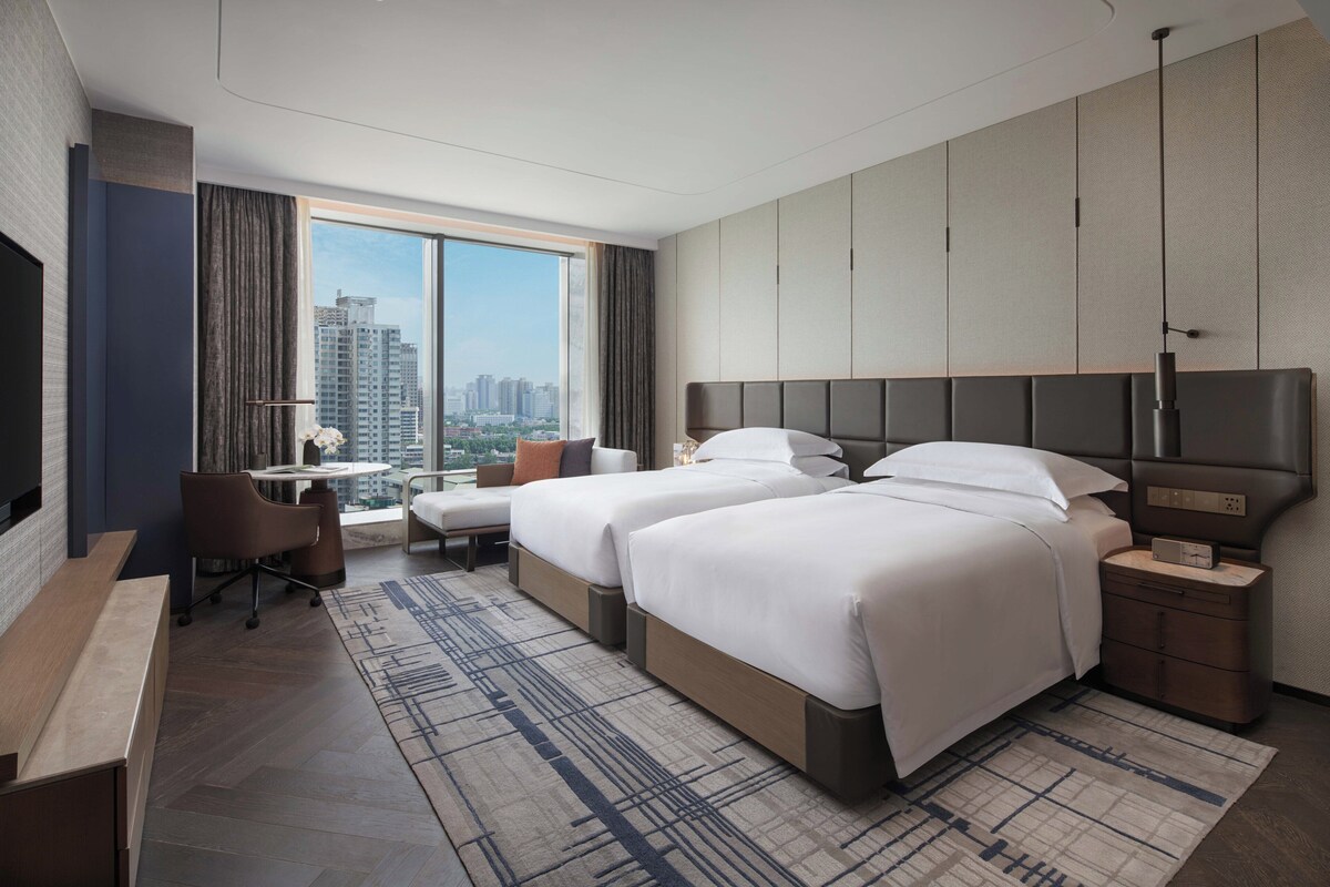 Una habitación de hotel moderna con dos camas, un escritorio, una silla y un ventanal con vistas a la ciudad.