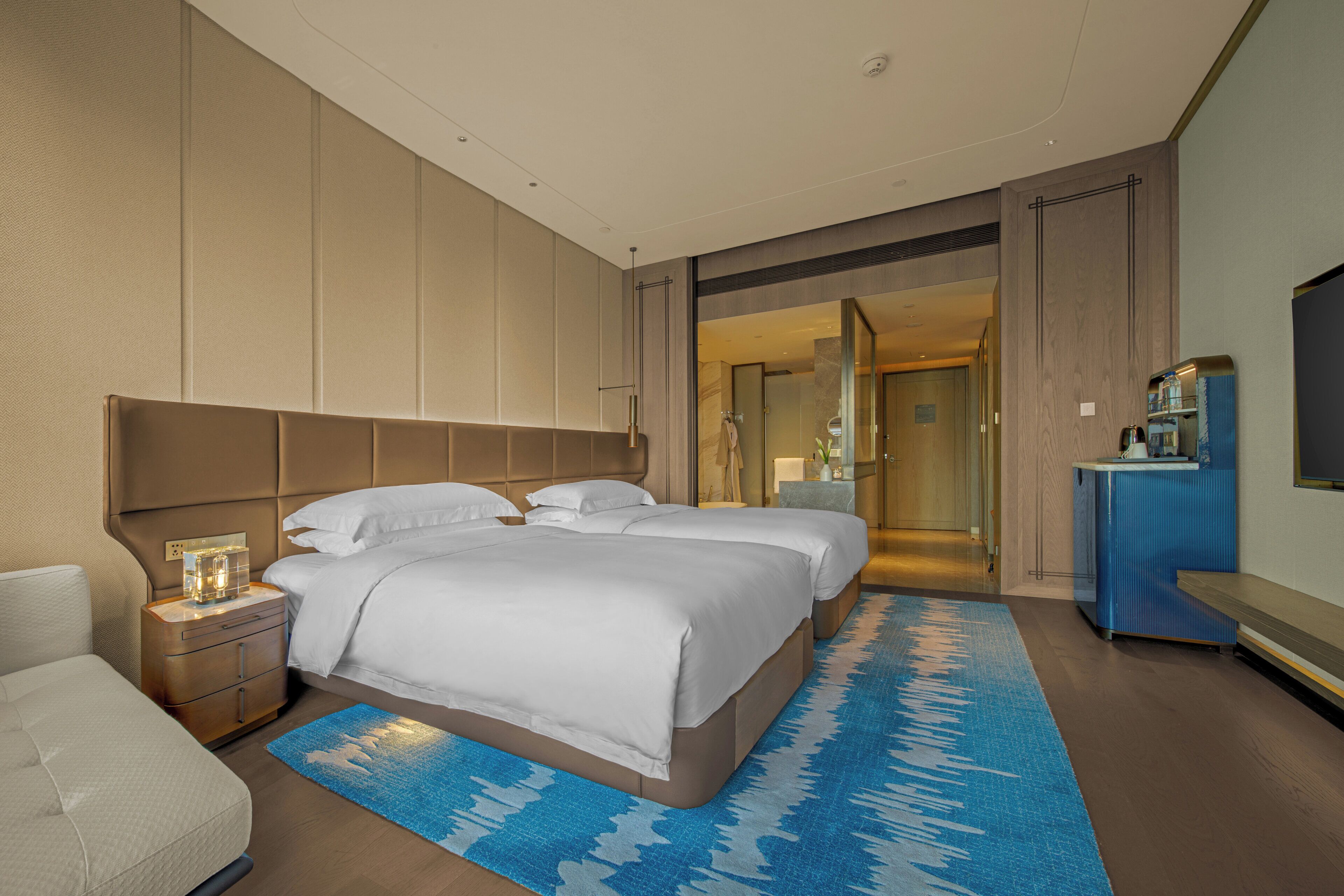 Une chambre d’hôtel moderne avec un grand lit, une table de chevet avec une lampe, une télévision et un tapis à motifs bleus.