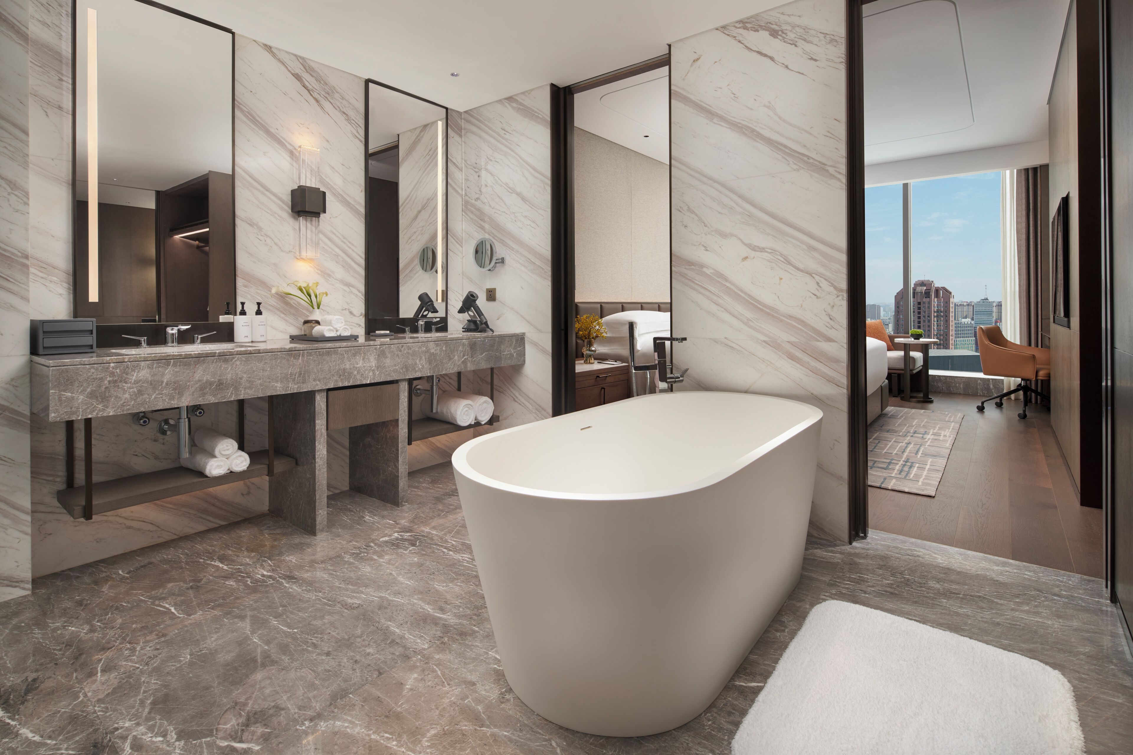 A modern bathroom with a freestanding bathtub, a large vanity with a mirror, and a city view through the window.
