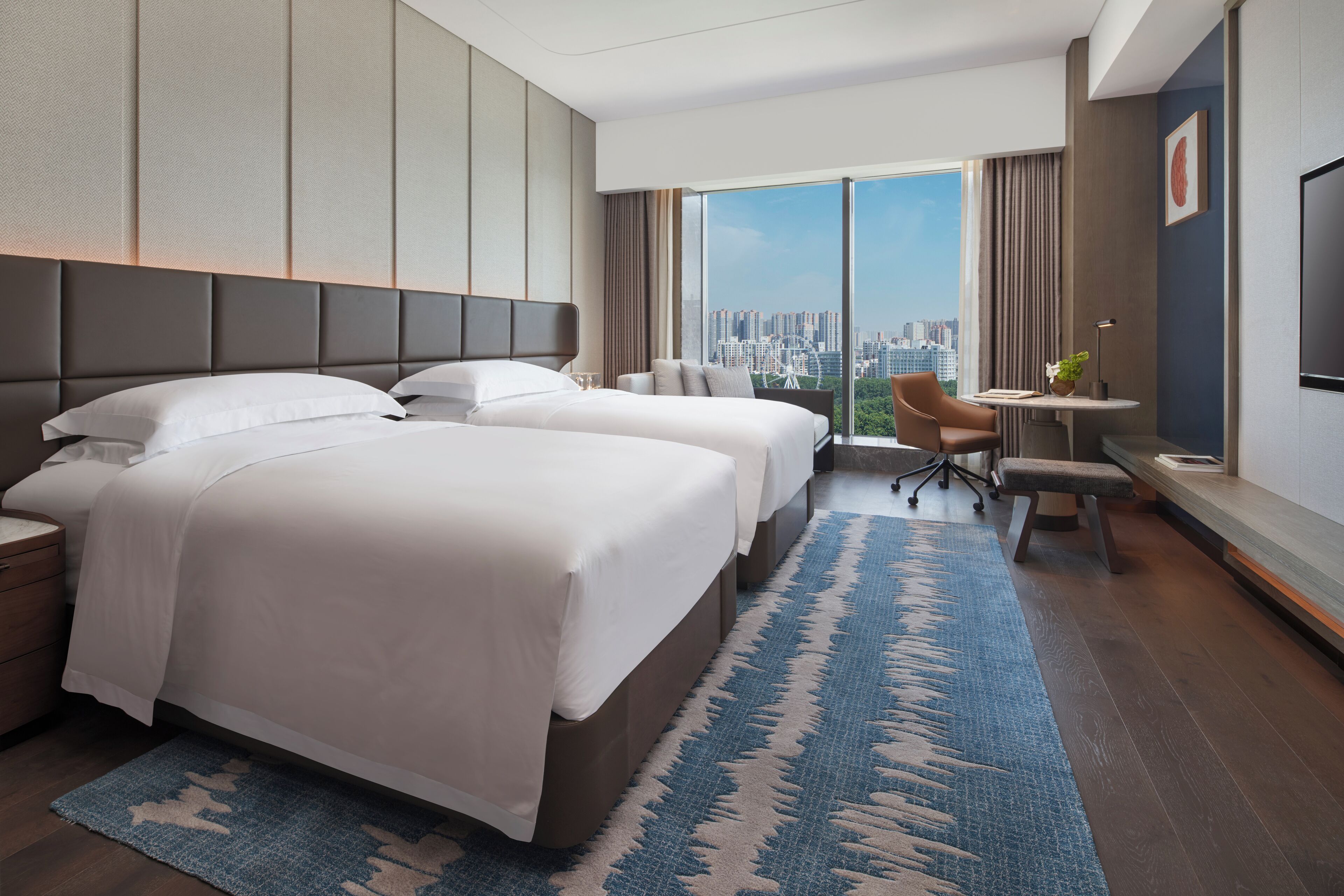 A hotel room with two beds, a desk, a chair, and a large window with a city view.