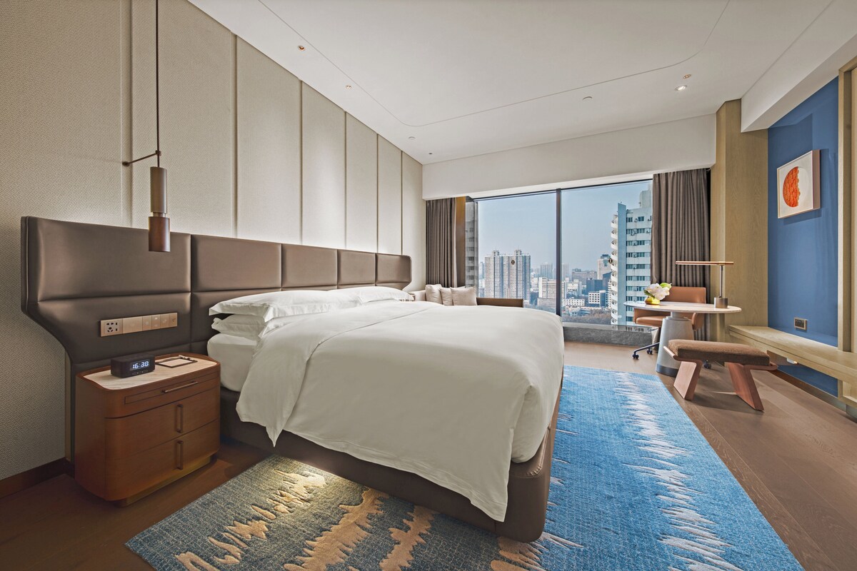 Habitación de hotel moderna con una cama grande, una mesita de noche, un escritorio y vistas a la ciudad.