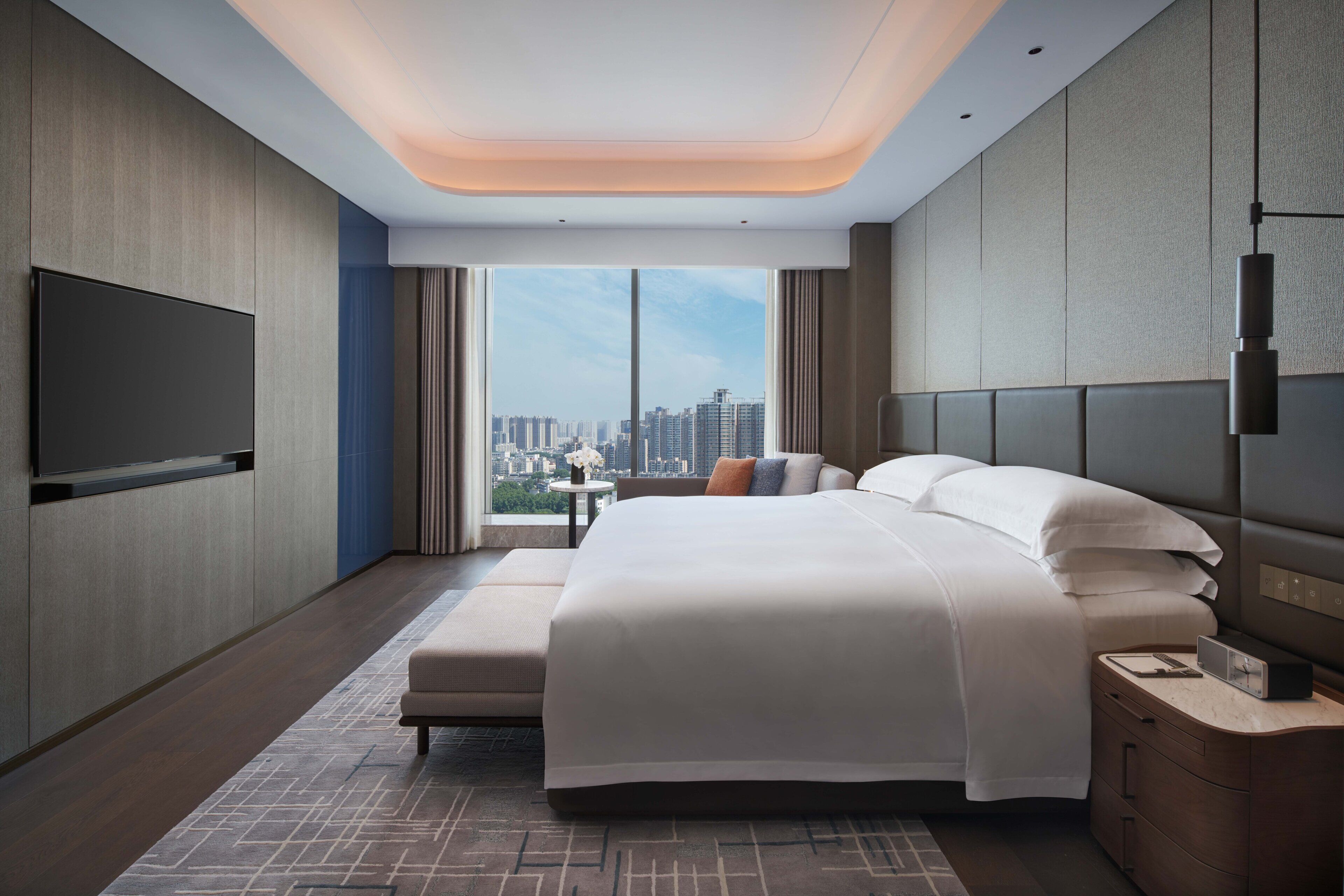 A modern hotel room with a large bed, a flat-screen TV, a bedside table, and a city view.