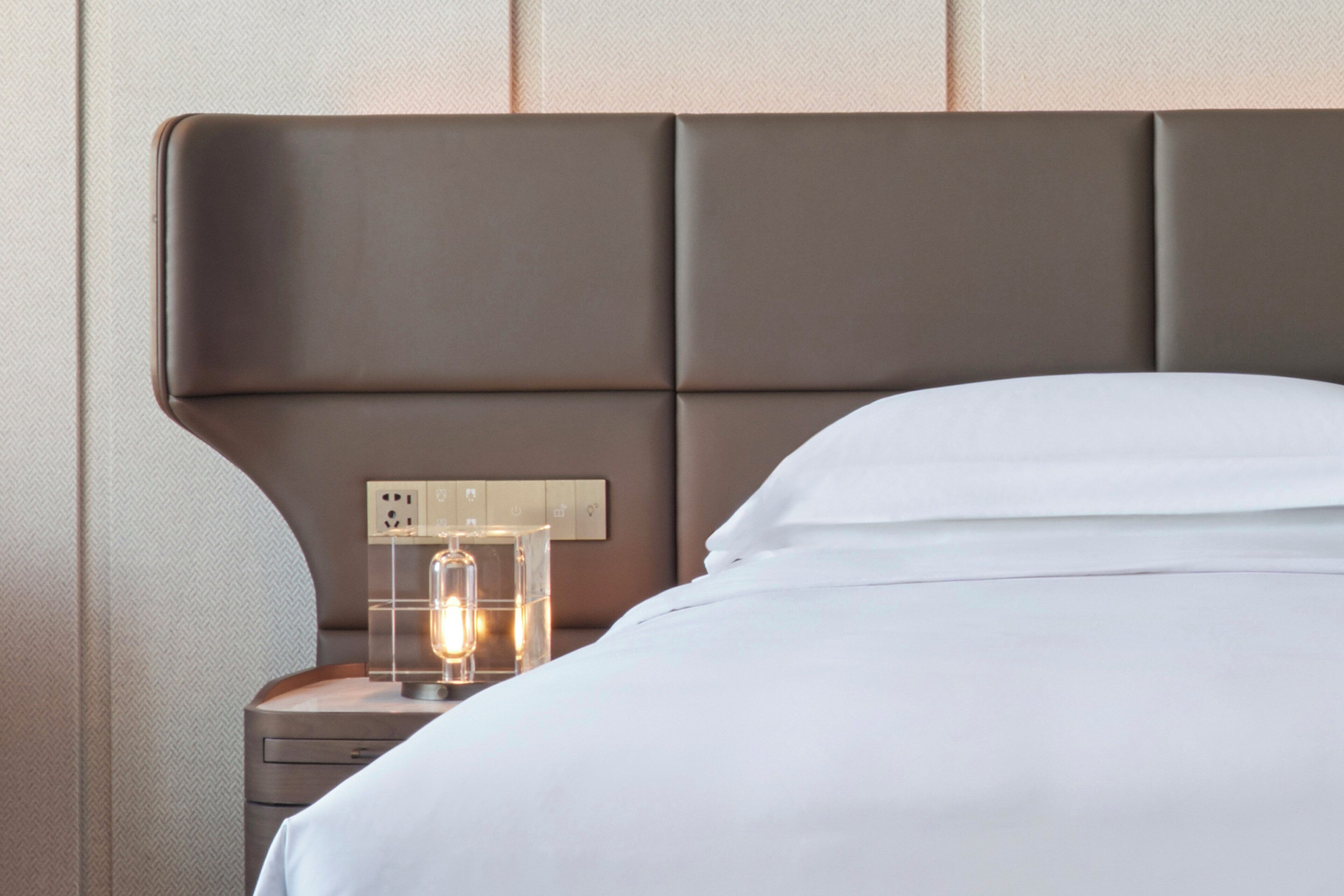 A neatly made bed with a headboard, a bedside table with a lamp, and a power outlet.
