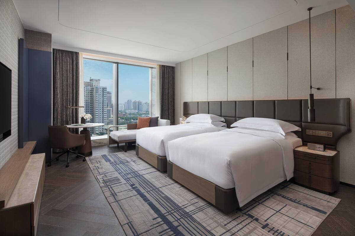 A modern hotel room with two beds, a desk, a chair, and a large window with a city view.