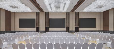 A large conference room with rows of white chairs, a large screen, and a chandelier.