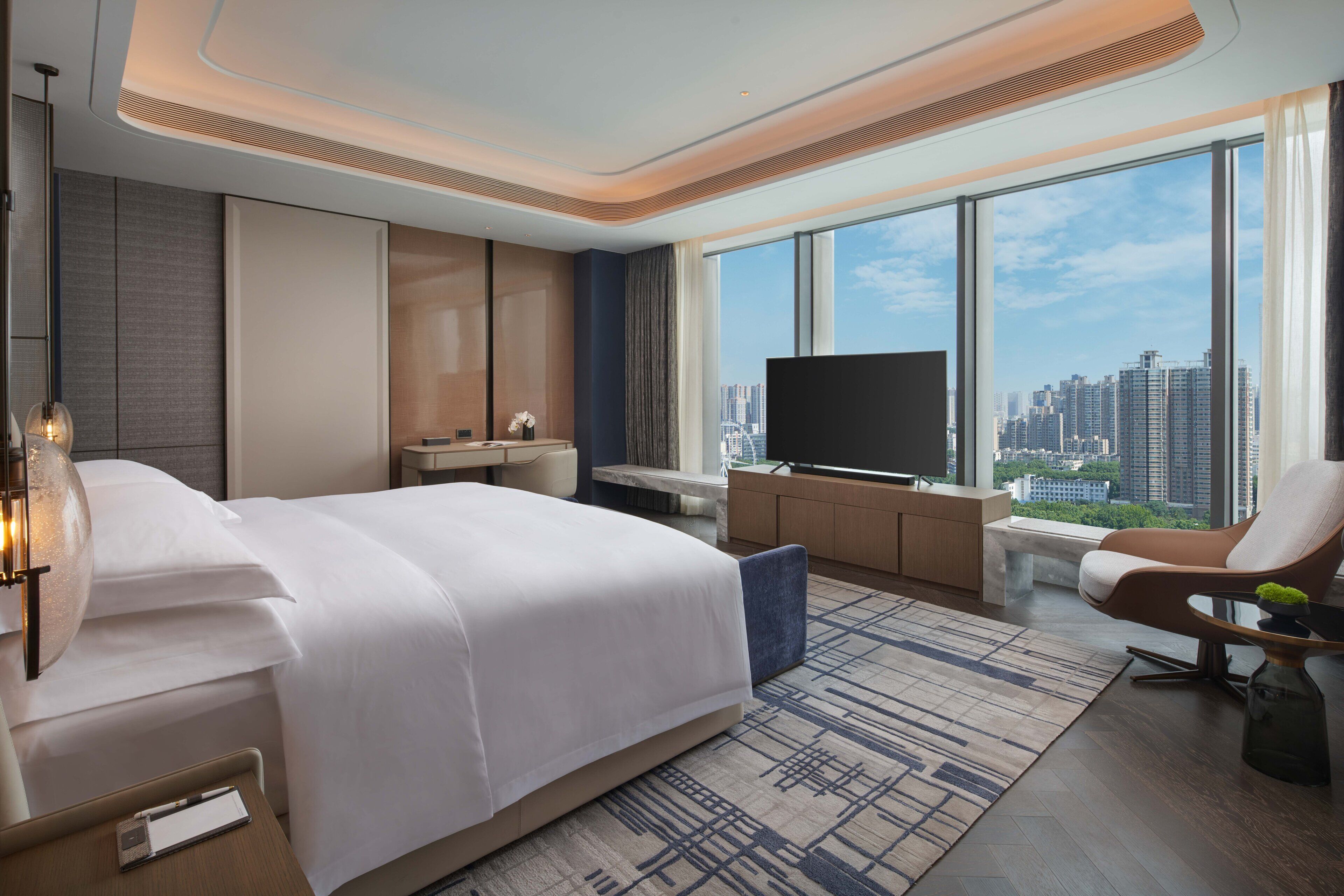 A modern hotel room with a large bed, a desk, a TV, and a view of the cityscape.
