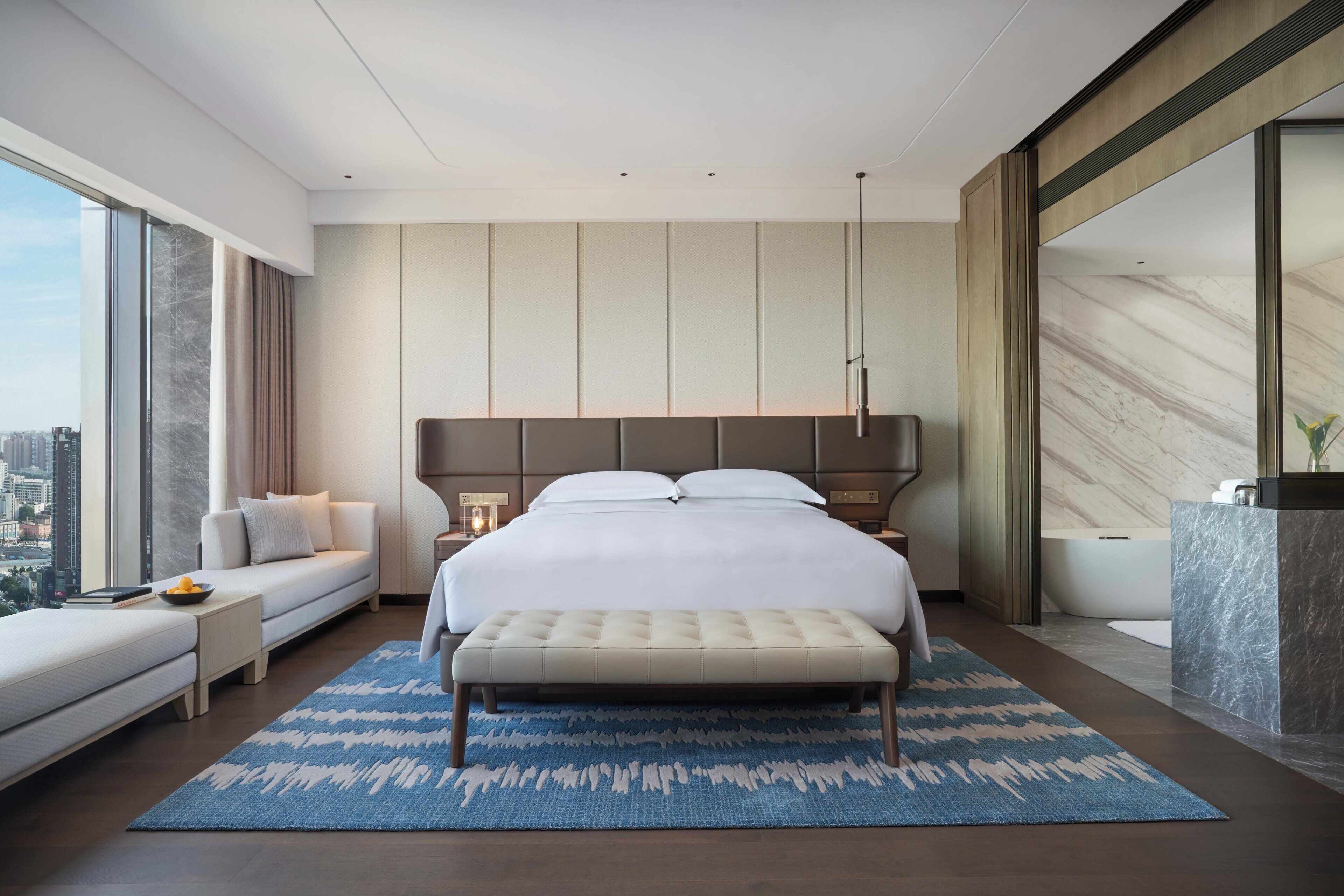 A modern hotel room with a large bed, a seating area, a view of the city, and a bathroom with a marble countertop.