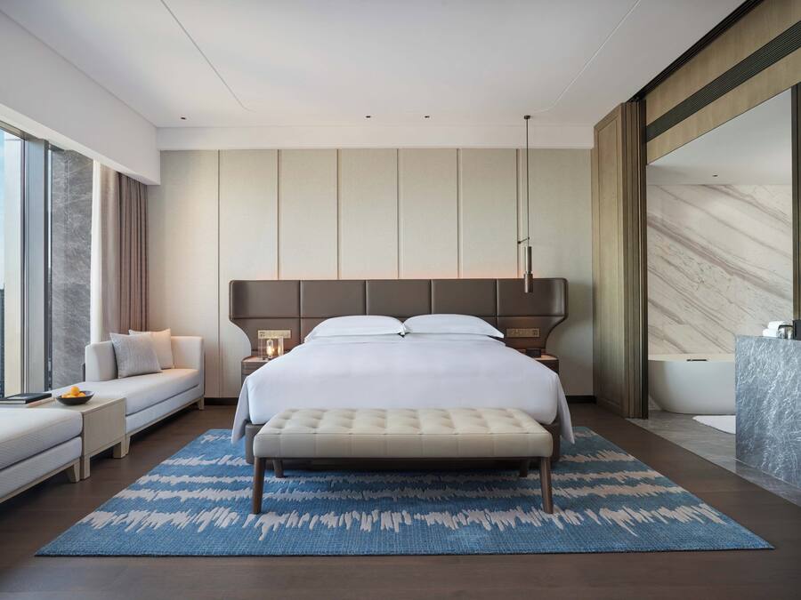 A modern hotel room with a large bed, a seating area, a view of the city, and a bathroom with a marble countertop.