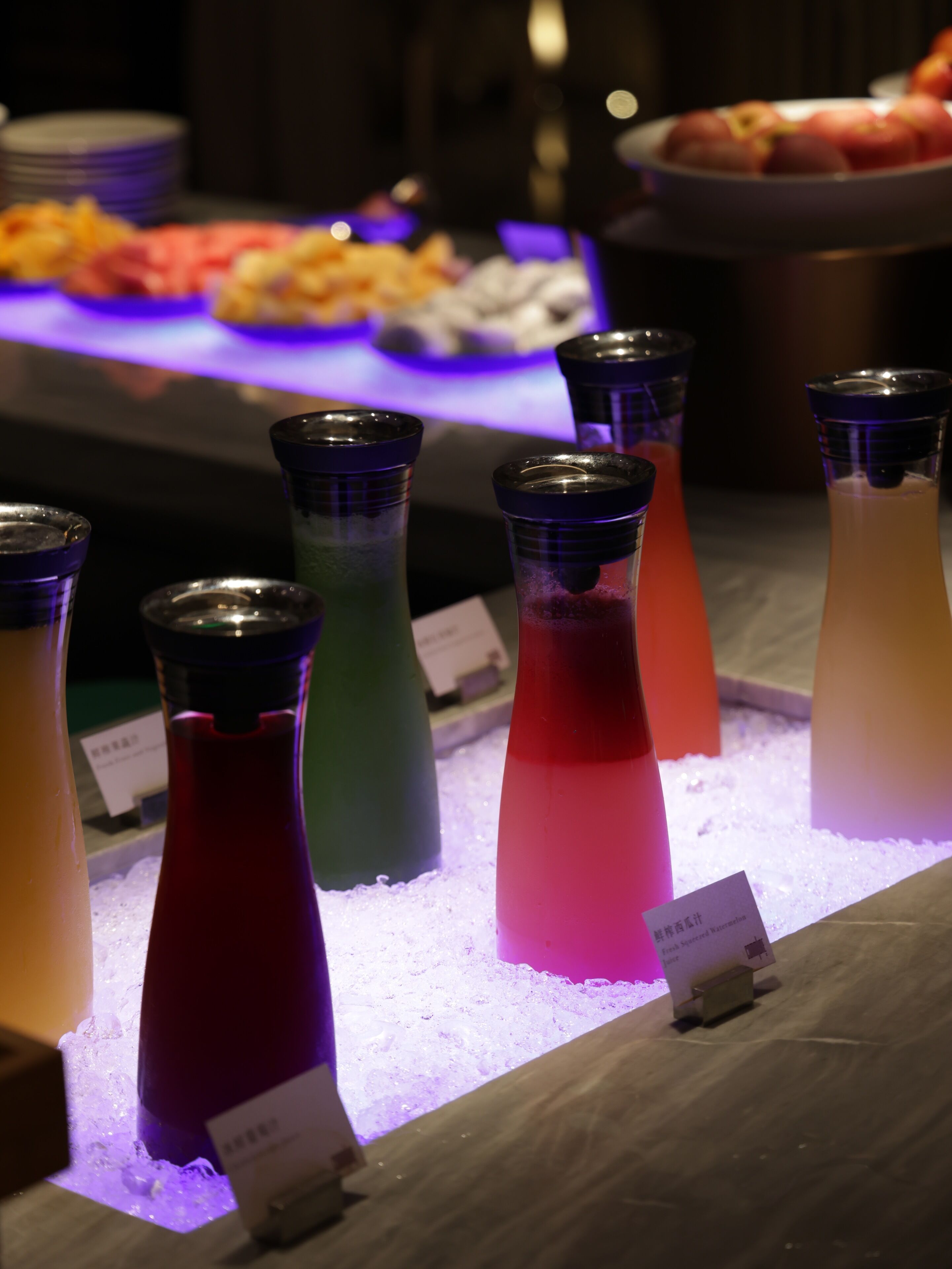 Variedad de bebidas coloridas en botellas de vidrio sobre un mostrador con fondo iluminado en púrpura.