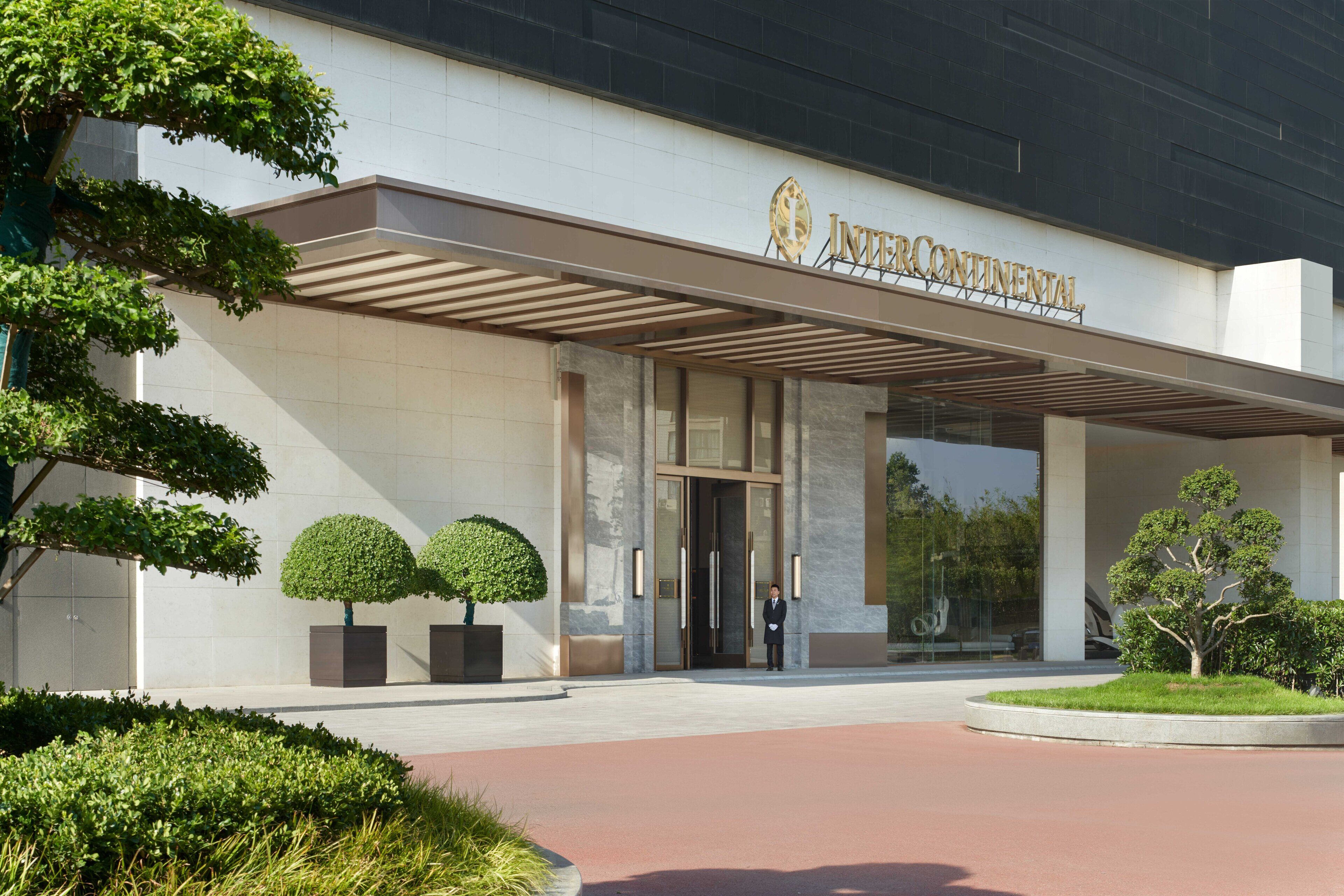 Ein moderner Hotelzugang mit einer überdachten Laube, einem Schild mit der Aufschrift 'INTERCONTINENTAL' und ordentlich geschnittenen Formschnitten.