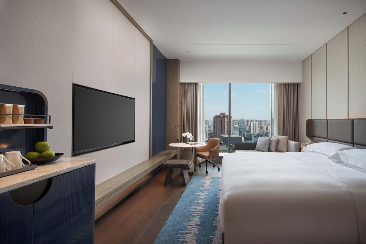 Habitación de hotel con una cama grande, un escritorio con televisor de pantalla plana, una pequeña mesa con una silla y vistas a la ciudad a través de amplios ventanales.