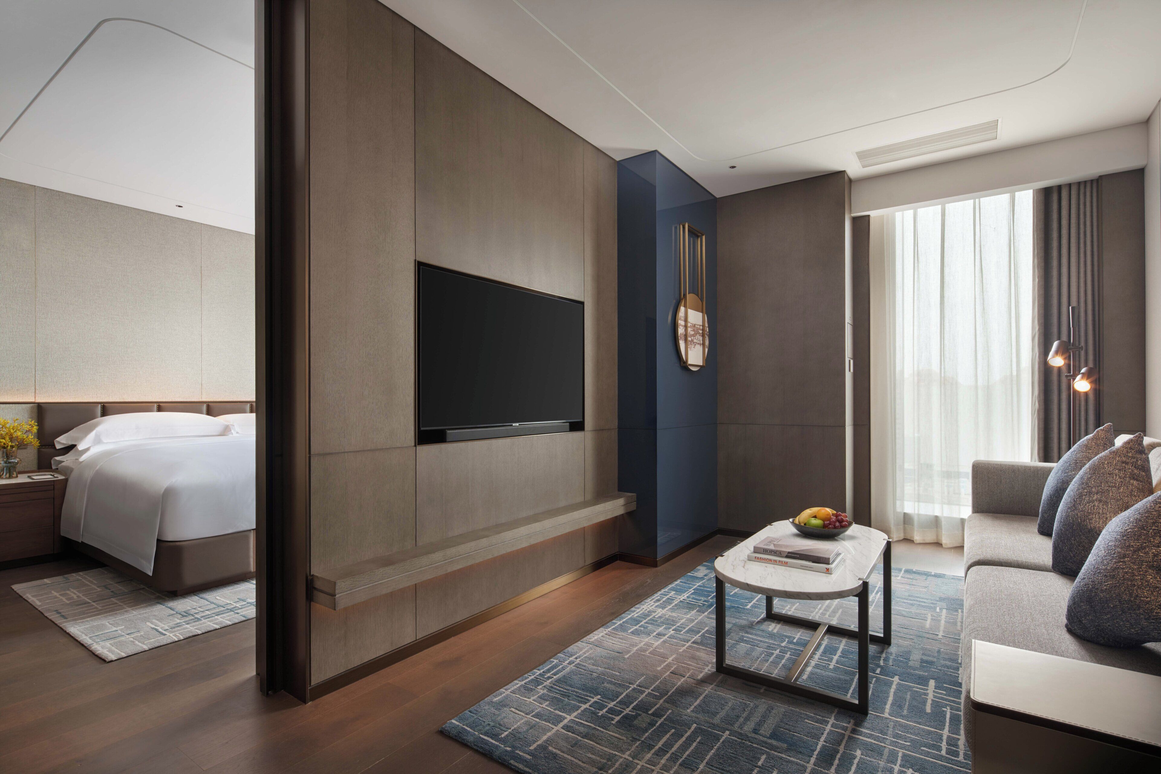 A modern hotel room with a bed, a flat-screen TV mounted on the wall, a coffee table with books, a sofa, and a large window with curtains.