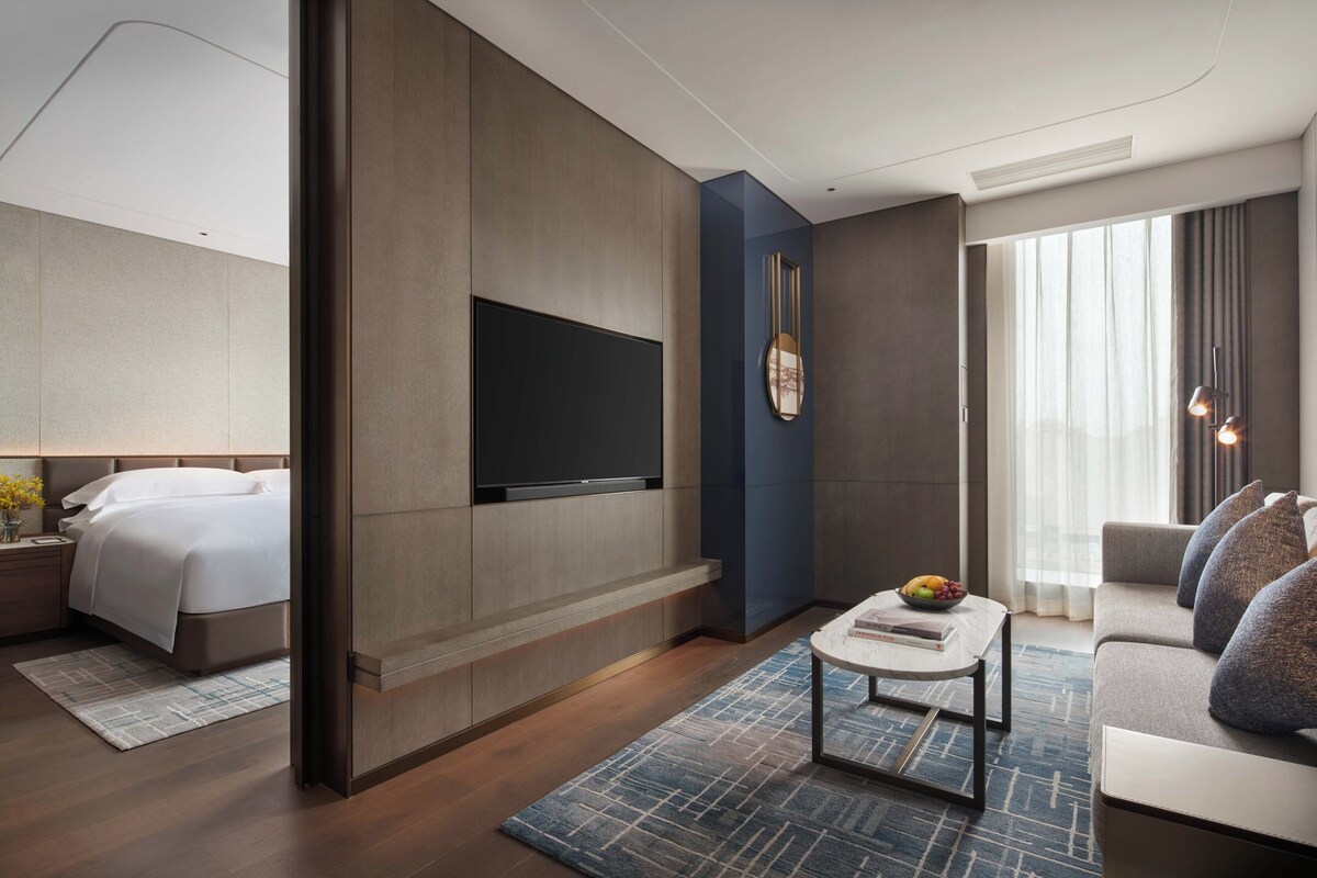 Una habitación de hotel moderna con cama, televisor de pantalla plana en la pared, sofá, mesa de centro y un ventanal con cortinas.