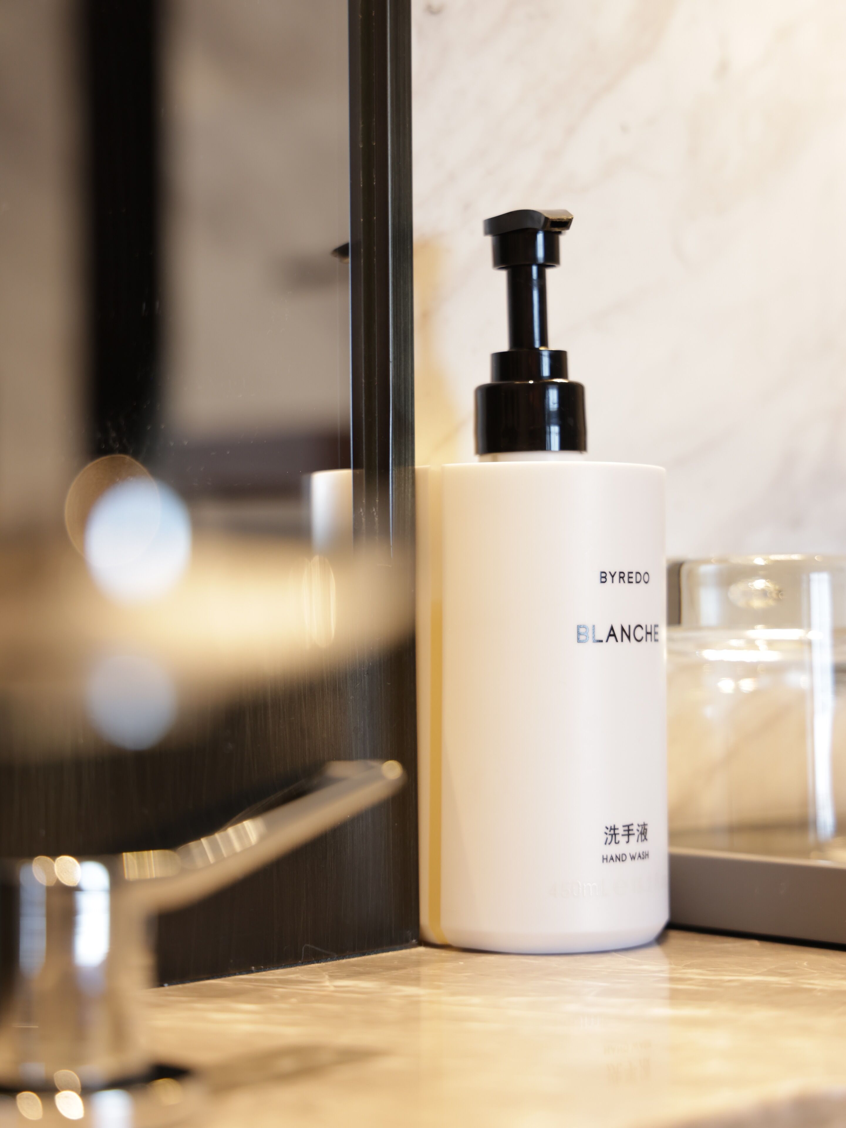 A white hand wash bottle with a black pump, labeled "BYREDO BLANCHE," placed on a marble countertop next to a glass of water.