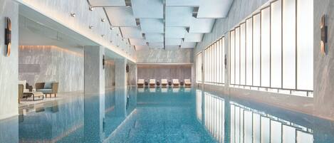 Una piscina interior moderna con superficie de agua azul cristalina, una zona de estar con sillones reclinables y un diseño de techo contemporáneo con iluminación empotrada.