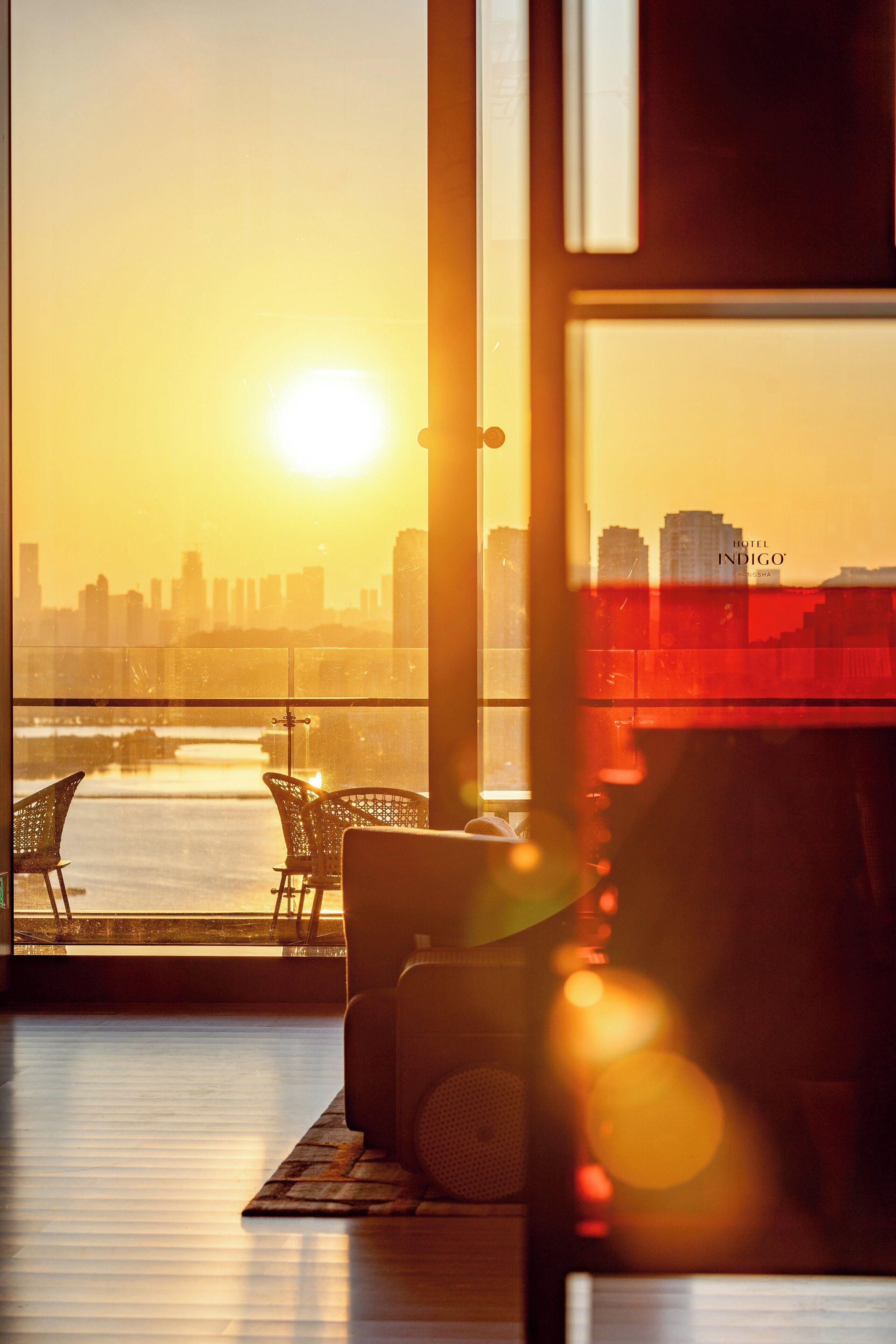 Une pièce offrant une vue sur les toits d’une ville au coucher du soleil, avec une grande fenêtre et un coin salon équipé de fauteuils.