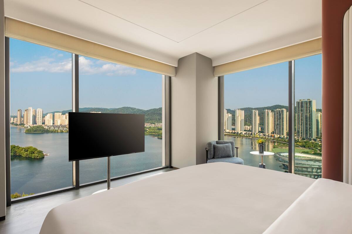 A modern hotel room with a large bed, a flat-screen TV, and a city view through large windows.