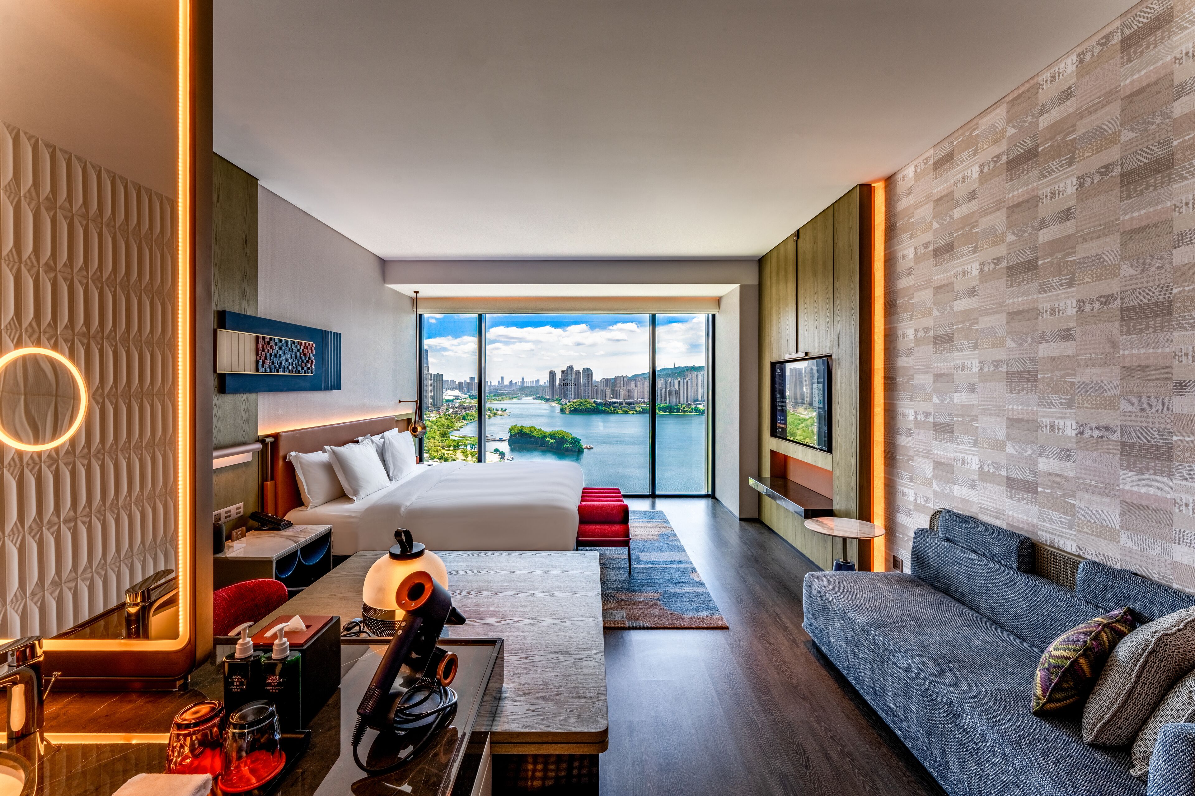 Habitación de hotel moderna con una cama grande, un sofá, un escritorio y vistas a un paisaje urbano y al río.