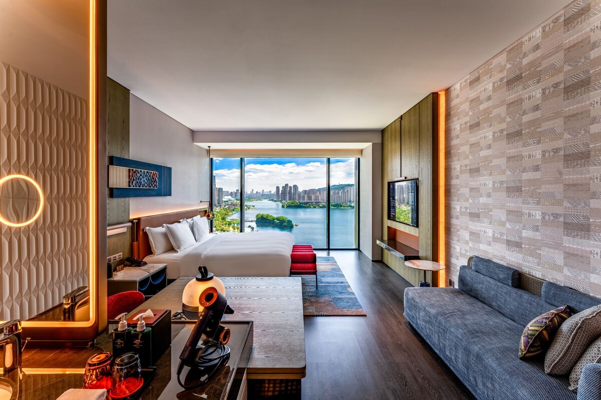 A modern hotel room with a large bed, a sofa, a desk, and a view of a cityscape and river.