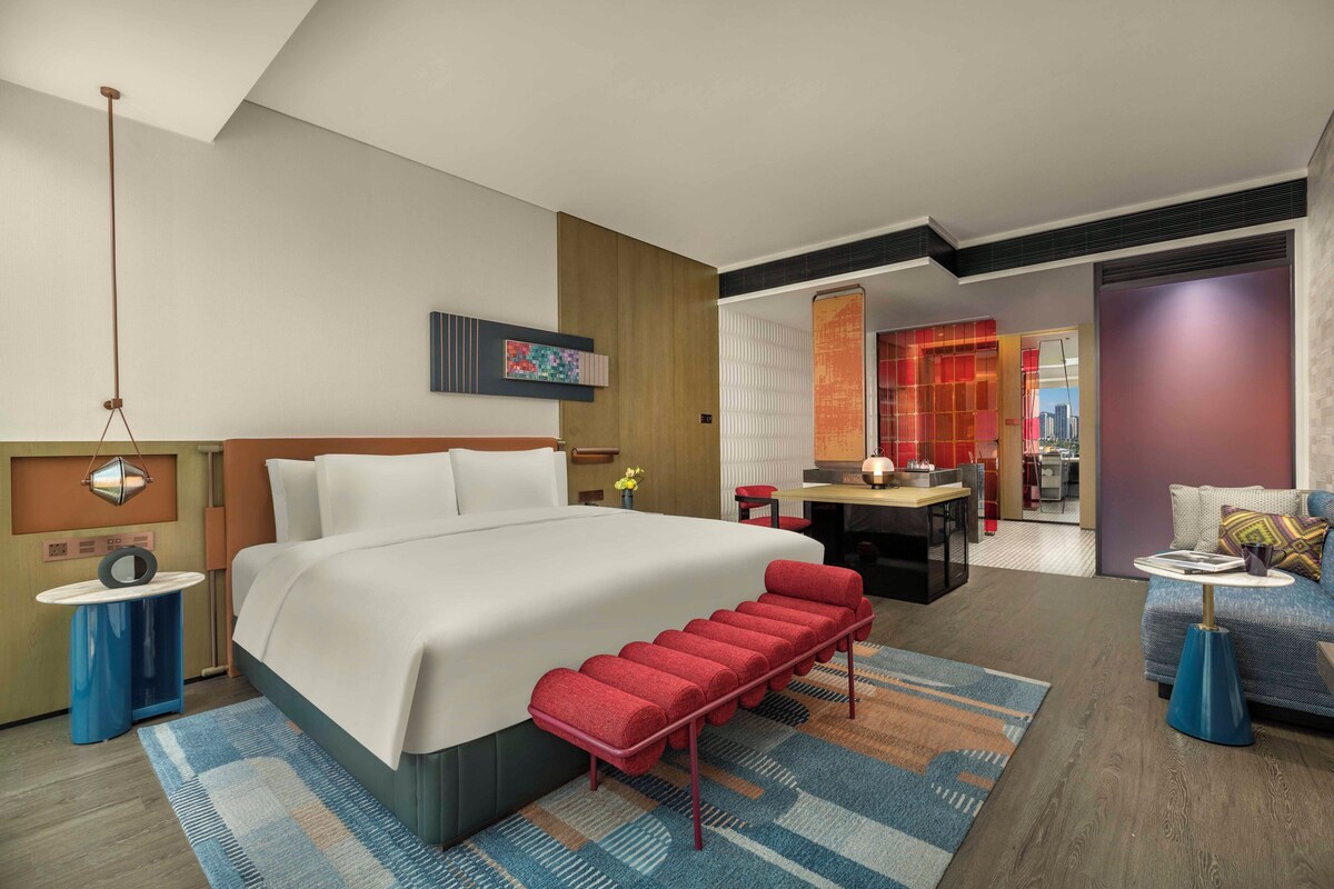 A modern hotel room with a large bed, a red bench, a sofa, a dining area, and a view of the city.