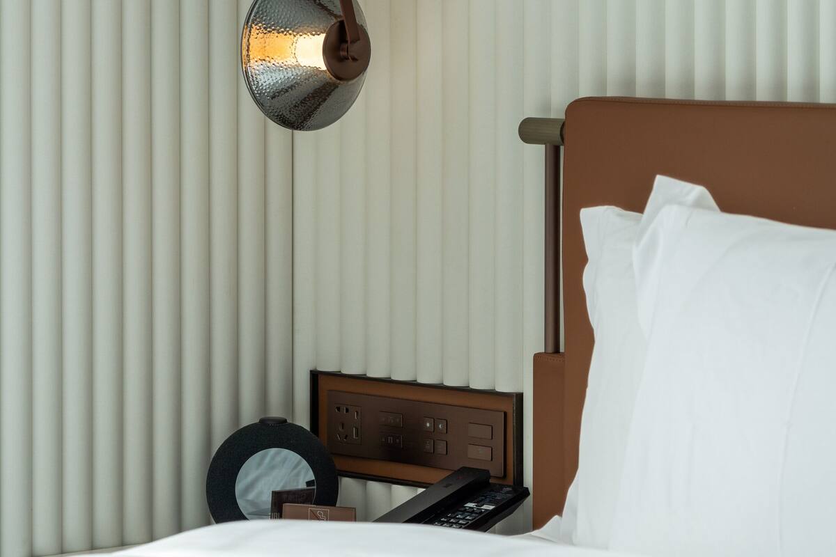 A hotel room with a bed, a bedside table with a lamp, and a window with blinds.