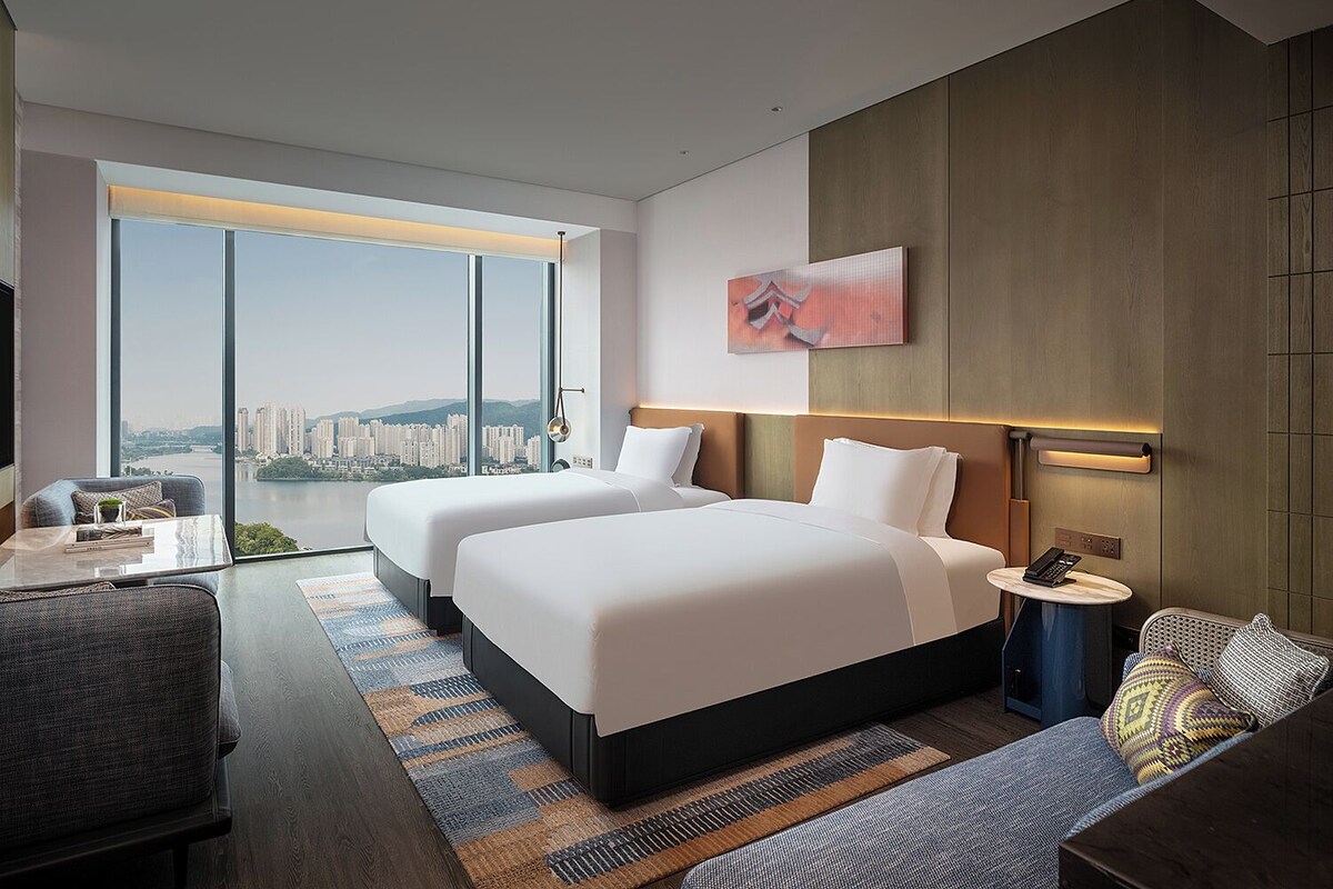 A modern hotel room with two beds, a large window offering a city view, a small table, and a sofa.
