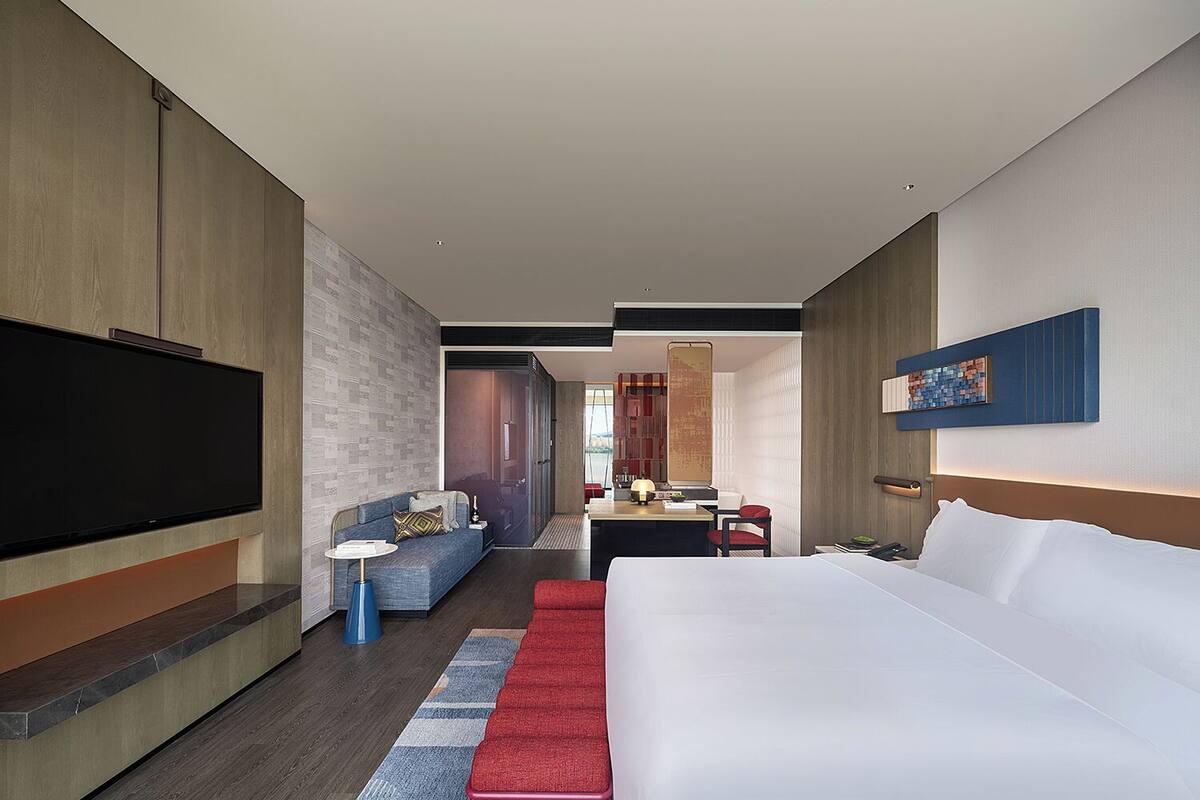 A modern hotel room with a large bed, a flat-screen TV, a seating area with a sofa and armchairs, and a dining table.
