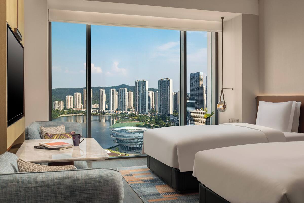 A hotel room with a large window offering a city view, two beds, a sofa, a coffee table, and a TV.