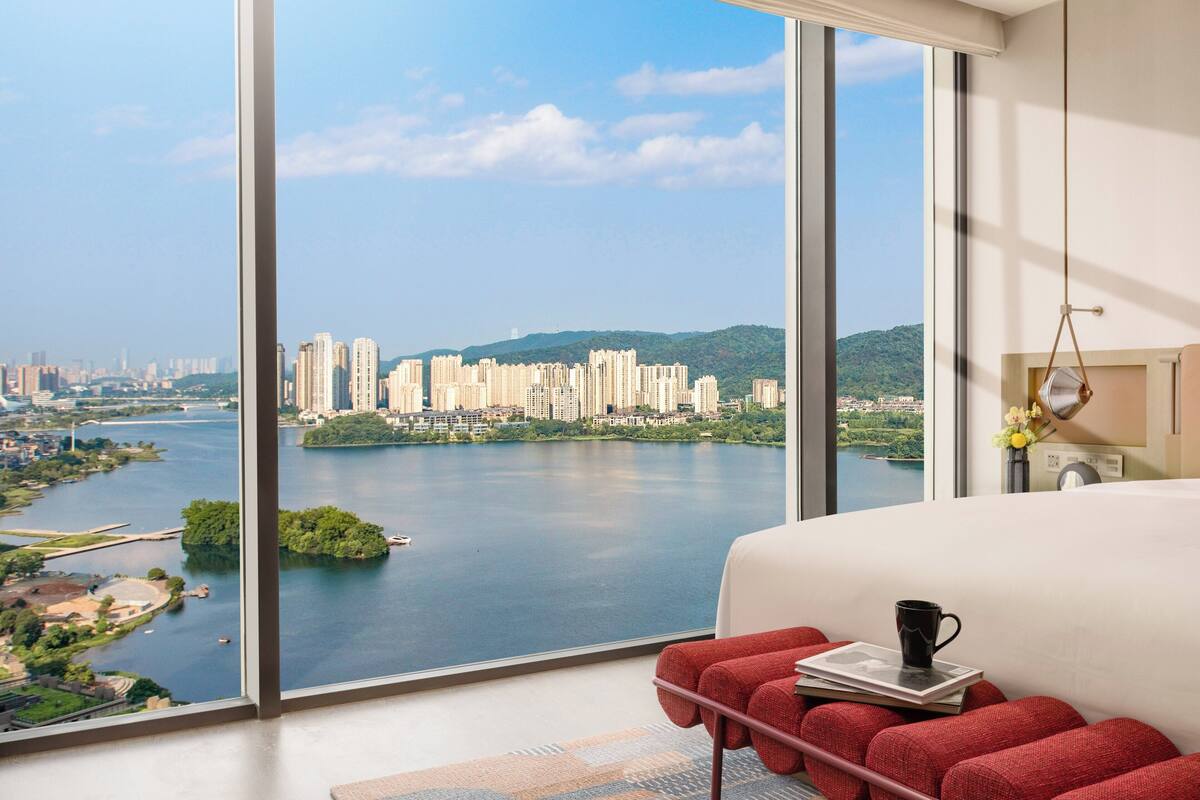 A modern hotel room with a large window offering a city view, a bed with a red headboard, and a small table with a book and a mug.
