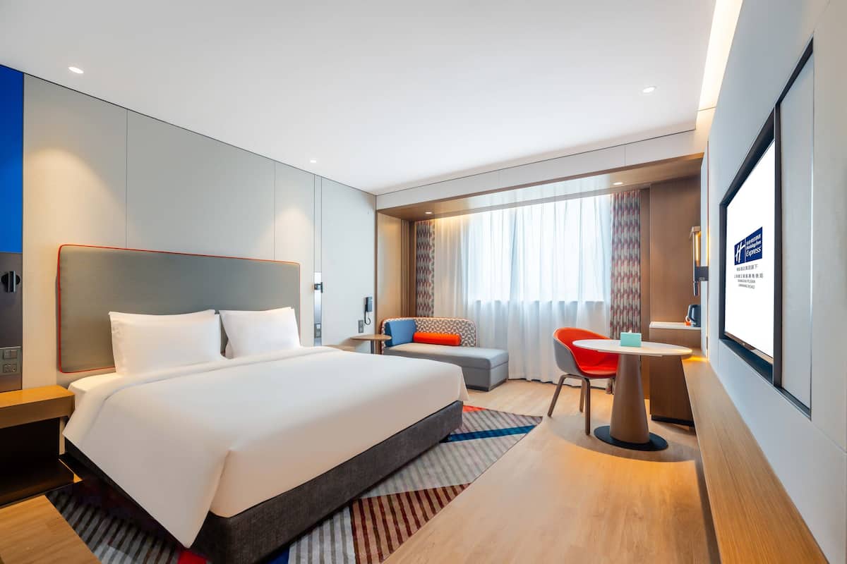 A modern hotel room with a large bed, a sofa, a desk, and a TV screen.