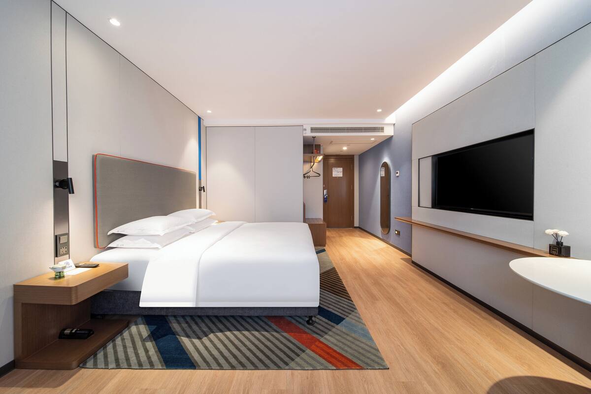 A modern hotel room with a large bed, a bedside table, a TV, and a bathroom with a sink and mirror.