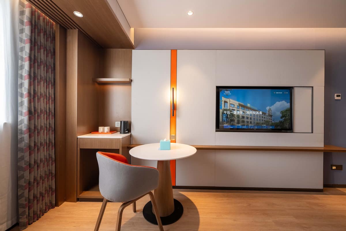 A modern hotel room with a round table, a single chair, a TV mounted on the wall, and a window with curtains.