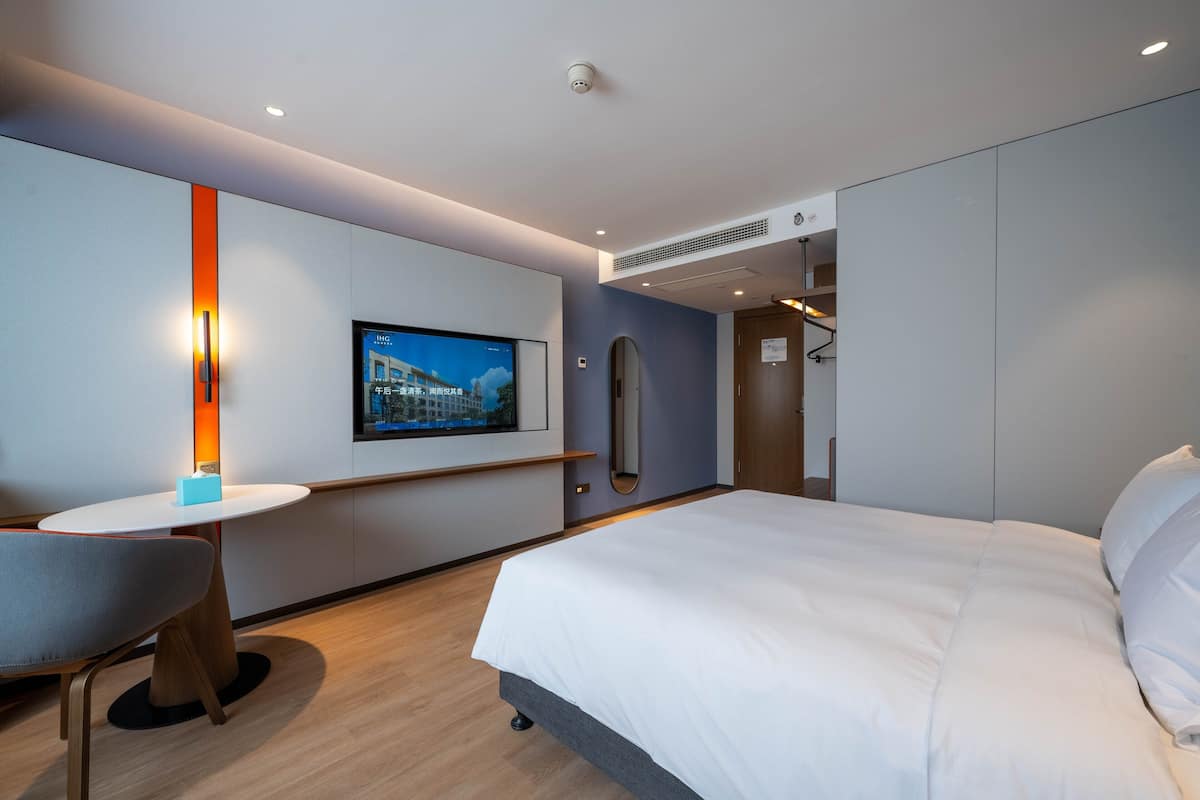 A modern hotel room with a large bed, a desk with a chair, a flat-screen TV mounted on the wall, and a view of the cityscape.