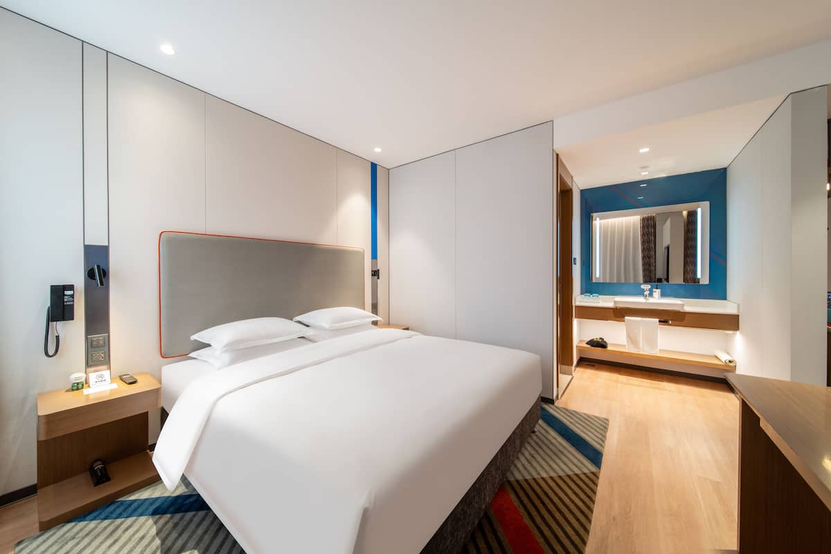 A modern hotel room with a large bed, a bedside table with a phone and a coffee cup, a bathroom with a mirror and sink, and a striped rug.