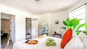 Un dormitorio ordenado con cama, una mesita de noche con una planta, un espejo y una ventana con luz natural.