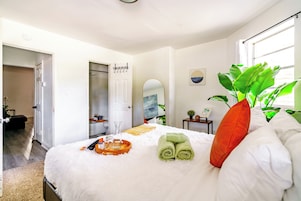 Un dormitorio ordenado con cama, una mesita de noche con una planta, un espejo y una ventana con luz natural.