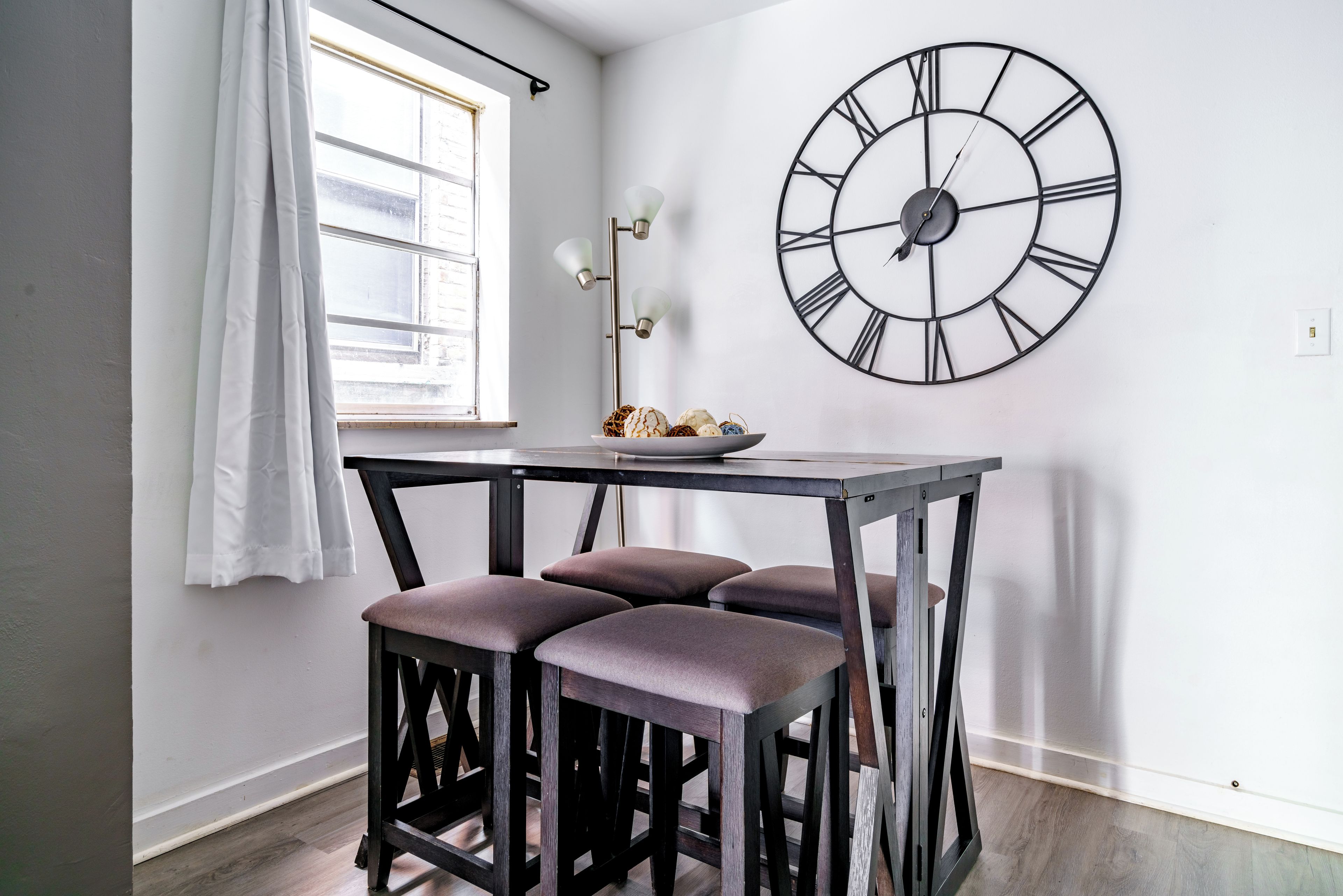 Une pièce comprenant une table, trois tabourets, une horloge murale, une fenêtre avec des rideaux et un lampadaire.