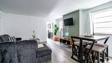 A modern living room with a grey sofa, a dining table with four chairs, a flat-screen TV mounted on the wall, and a potted plant.