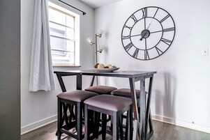 A room with a table, three stools, a wall clock, a window with curtains, and a floor lamp.