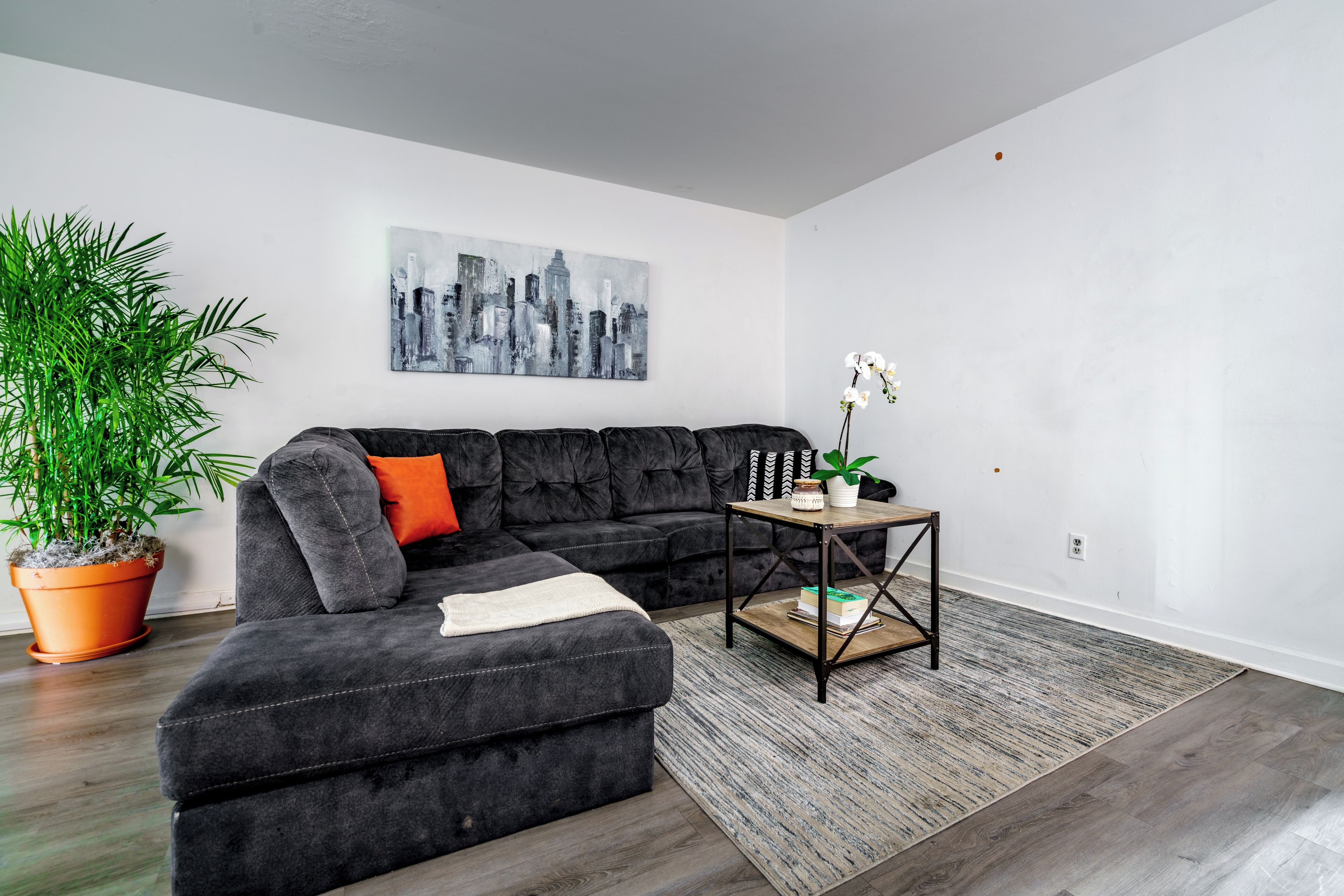 A modern living room with a sectional sofa, a cityscape painting, a potted plant, and a small table with books.