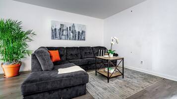 A modern living room with a sectional sofa, a cityscape painting, a potted plant, and a small table with books.
