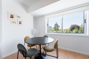 Una habitación moderna con una mesa redonda, cuatro sillas, un ventilador de pie y un ventanal con vistas a edificios y árboles.
