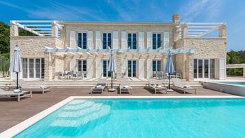 Un grande edificio in pietra con piscina, numerosi lettini e ombrelloni.