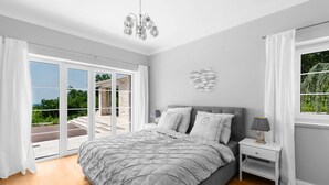 Ein Schlafzimmer mit einem großen Bett, einem Nachttisch, einer Lampe und Blick ins Freie durch eine Glastür.