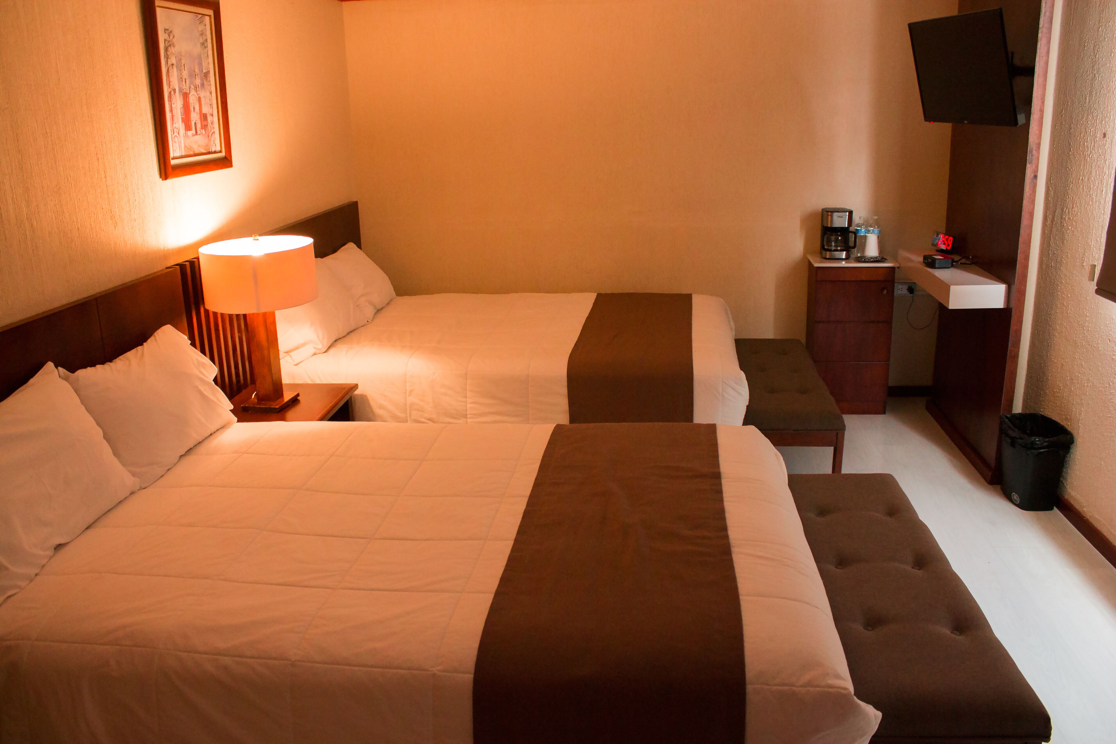 A hotel room with two beds, a bedside table with a lamp, a small desk with a coffee maker, and a trash bin.